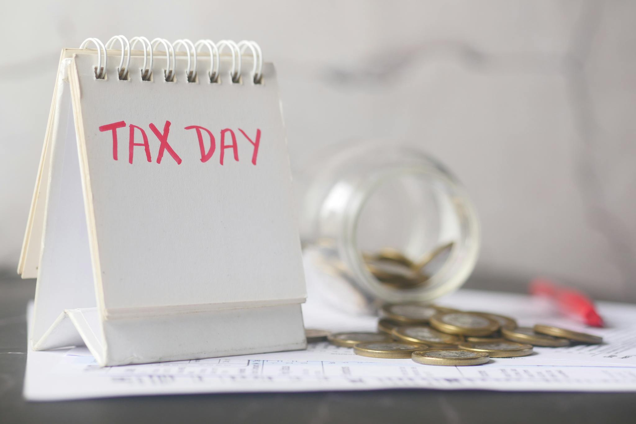 A close-up of a table calendar marked 'Tax Day' with a jar of coins and notes, symbolizing financial planning.